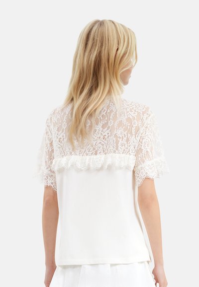 Blondebluse med korte, flæseærmer, prydet med et hvidt blondeoverlay og en ensfarvet hvid ryg. Blød tekstur og delikat blomstermønster.