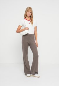 T-shirt blanc à manches courtes avec des accents rouges, pantalon large en tricot gris, et un sac à bandoulière blanc. Modèle portant des sneakers blancs.