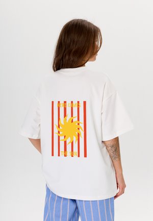 Witte oversized t-shirt met een gele zonne-afbeelding en rode tekst "HERE COMES THE SUN" en verticale rode strepen op de achterkant.