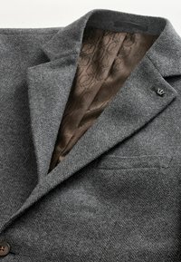 Blazer en laine gris à chevrons avec doublure intérieure marron à motifs, col cranté et petite épingle métallique sur le col.