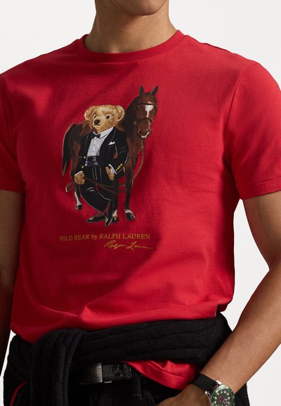 Κόκκινο βαμβακερό μπλουζάκι με σχέδιο ενός αρκούδου σε τοξοτο παντελόνι δίπλα σε ένα άλογο, διακοσμημένο με μαύρες και χρυσές λεπτομέρειες. Περιλαμβάνει κείμενο "Polo Bear by Ralph Lauren".