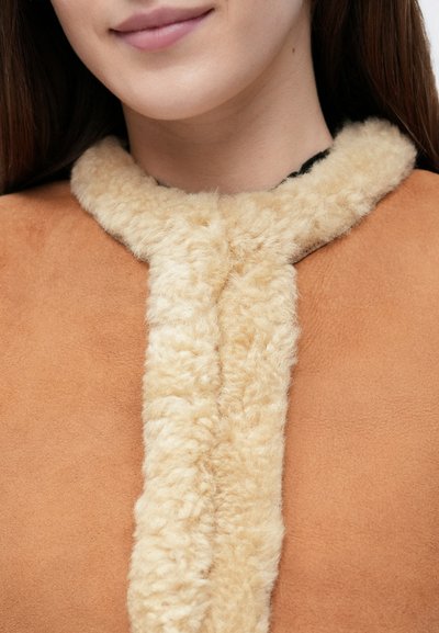 Κοντινό πλάνο ενός χαμογελαστού ατόμου που φοράει ένα καφέ παλτό με χοντρή, κρεμ επένδυση shearling γύρω από το λαιμό και το μπροστινό άνοιγμα.