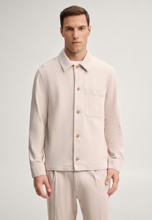 Uomo in piedi rivolto frontalmente che indossa una camicia beige abbottonata con una sola tasca sul petto e pantaloni beige abbinati, su uno sfondo chiaro e uniforme.