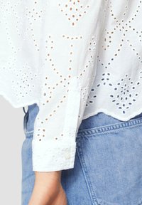 Witte katoenen blouse met uitgesneden borduursel en gegolfde zoom. Lange mouwen met knoopmanchetten. Gedragen over lichtblauwe denim.