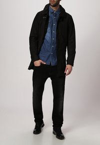 Svart lång kappa över en blå jeansskjorta, tillsammans med svarta jeans och mörka skor. Funktioner inkluderar en krage, framsida med dragkedja, och fickor.