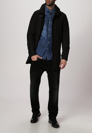 Abrigo largo negro sobre una camisa de mezclilla azul, combinado con jeans negros y zapatos oscuros. Las características incluyen un cuello, cierre frontal y bolsillos.