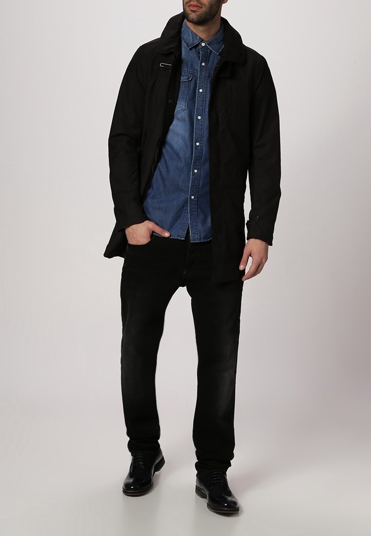 Svart lång kappa över en blå jeansskjorta, tillsammans med svarta jeans och mörka skor. Funktioner inkluderar en krage, framsida med dragkedja, och fickor.