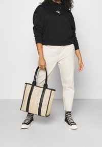 Bolso tote beige con acentos negros, fabricado en un material texturizado. Presenta dos asas robustas y rayas verticales. Persona vestida con pantalones claros y sudadera negra.