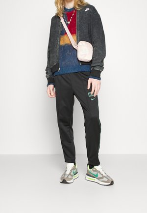 Giacca bomber Nike in denim nero, maglione a maglia multicolore, pantaloni della tuta neri con accenti verdi, sneakers grigie con dettagli verdi e borsa rosa.