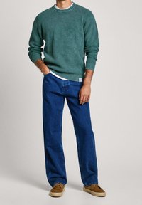 Grüner Strickpullover mit strukturiertem Muster, kombiniert mit lockeren blauen Jeans und braunen Schuhen, mit einem weißen Innenkragen.