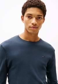 Granatowy, długi rękaw, koszulka z okrągłym dekoltem wykonana z teksturowanej tkaniny, z małym haftowanym logo na lewej piersi. Widoczne są przeszycia.