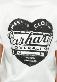 Camiseta blanca con un diseño gráfico negro en forma de corazón y el texto "Carhartt Overalls Work in Progress" en estilo vintage.