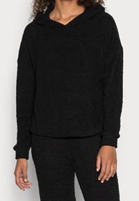 Schwarze, flauschige Kapuzenjacke aus weichem Material, mit lockerer Passform, langen Ärmeln und einer Kapuze. Dazu passende Unterteile.