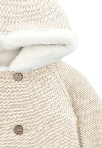 Chaqueta de punto beige con capucha forrada de pelusa blanca, costuras reforzadas y dos botones redondos marrones en el frente. Textura suave en toda la prenda.