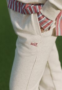 Jasnobeżowe spodnie dresowe o fakturowanym wykończeniu, z małym czerwonym haftem "self." przy kieszeni oraz widoczną wzorzystą koszulą.