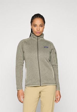 Vrouw met gevlochten haar, gekleed in een grijze fleece jas met rits en beige broek, staand voor een effen witte achtergrond.