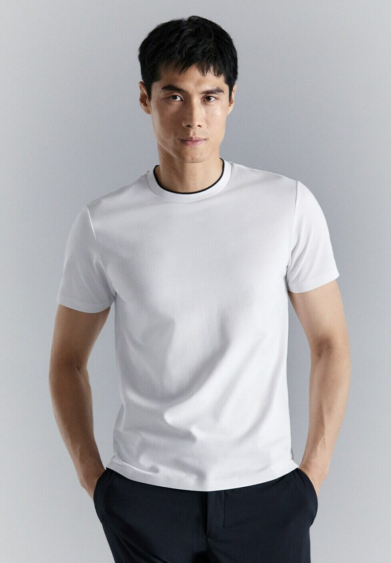 Homme aux cheveux courts noirs portant une chemise blanche à manches courtes unie et un pantalon foncé, debout les mains dans les poches devant un fond gris.