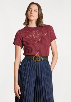 Femme portant un haut en maille bordeaux à manches courtes avec des trous décoratifs, une ceinture noire à boucle dorée, et un pantalon plissé taille haute bleu marine.
