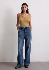 Vrouw met donker haar draagt een mouwloos beige topje, hoge taille wijdvallende blauwe jeans en bruine schoenen, staande tegen een effen lichte muur.