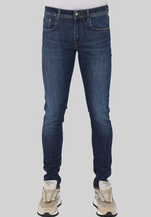 Uomo che indossa jeans blu scuro aderenti e sneakers beige in piedi davanti a uno sfondo grigio neutro.