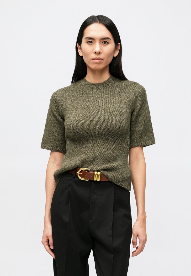 Olivovo zelený pletený sveter s krátkym rukávom a okrúhlym výstrihom, skombinovaný s čiernymi nohavicami s vysokým pásom a hnedým opaskom so zlatou prackou.