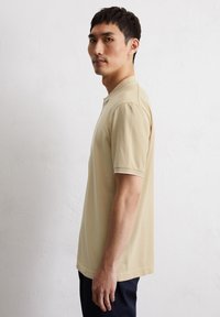 Beżowa koszulka polo z krótkim rękawem, z kołnierzykiem i białym wykończeniem, uszyta z miękkiego materiału. Model przedstawiony z profilu na jasnym tle.