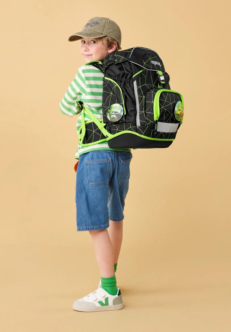 Zaino nero con motivo geometrico verde, diverse tasche, accessori grigi e dettagli riflettenti. Indossato da un bambino con una maglietta a righe e pantaloni corti.