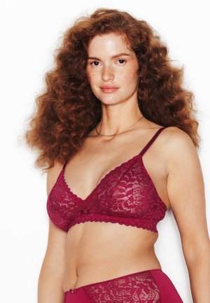 Donna dai capelli rossi con lentiggini che indossa un reggiseno a bralette di pizzo bordeaux abbinato e biancheria intima a vita alta su sfondo bianco.