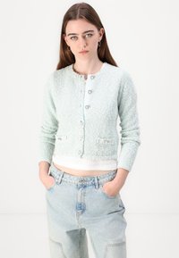 Light mint green cardigan with textured fabric, round neckline, and decorative buttons. Styled over a white top, paired with light denim jeans.