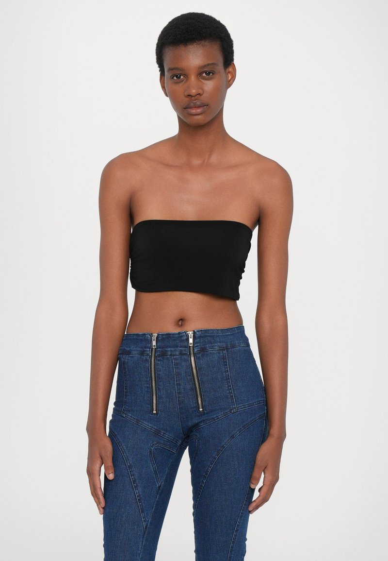Svart axelbandslös topp i kombination med mörkblå högmidjade jeans som har dubbla dragkedjor och vinklade sömdetaljer. Slät textur.