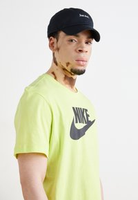 Camiseta Nike de color verde neón con logo negro, combinada con una gorra negra que tiene texto blanco. El sujeto tiene poco vello facial y variaciones en la piel.