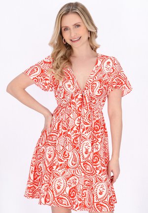 Glimlachende vrouw met golvend blond haar draagt een rood-witte, bloemenprint jurk met korte mouwen en knielengte, een V-hals en heeft haar hand op haar heup.