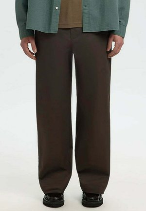 Uomo che indossa pantaloni larghi marrone scuro, scarpe nere, una camicia marrone e una camicia verde con bottoni, in piedi davanti a uno sfondo neutro.