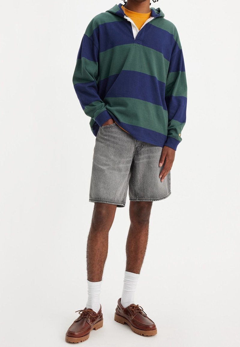 Personne portant un sweat à capuche oversize rayé bleu et vert, un short en jean gris, des chaussettes blanches et des chaussures en cuir marron, debout avec une main dans la poche.
