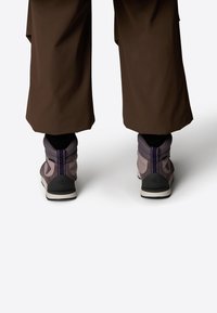 Botas de senderismo grises y moradas con un cuello acolchado y suelas negras, combinadas con pantalones sueltos marrones, vistas desde la parte trasera.