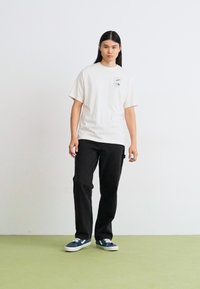 Hvid bomulds T-shirt med kortærmer og en brystlomme. Sorte bukser og blå sneakers. Ensfarvet baggrund med grøn gulv.