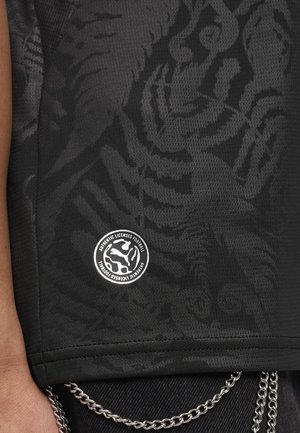 Czarna siateczkowa tkanina z subtelnym wzorem w paski tygrysa oraz białe okrągłe logo "Autentyczna Licencjonowana Piłka Nożna" nad srebrnym łańcuszkiem na czarnych spodniach.