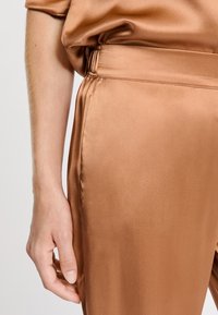 Braune Satin-Hosen mit glatter Textur, featuring elastischem Taillenbund und seitlichen Taschen, gezeigt neben einer teilweise sichtbaren Hand.