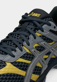 Κοντινό πλάνο μαύρου και κίτρινου παπουτσιού τρεξίματος ASICS με επάνω μέρος από πλέγμα, γκρι επικαλύψεις και μαύρα κορδόνια δεμένα πάνω σε λευκό φόντο.