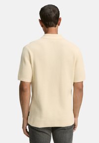 Mann mit kurzem dunklem Haar, der ein cremefarbenes, kurzärmliges, strukturiertes Strickhemd und dunkle graue Jeans trägt, von hinten gesehen.