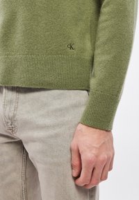 Zöld kötött pulóver bordázott mandzsettával és szegéllyel, finom logó részlettel. Világosszürke farmerrel párosítva. Lágy textúra.