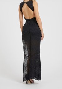 Robe maxi en dentelle noire avec motif en chevron, design à dos ouvert et silhouette ajustée. Présente des bretelles fines et une texture transparente.