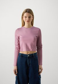 Roze duguljasti top s dugim rukavima, od glatkog materijala, uparen s hlačama od tamnog denima visoko strukiranim, s bočnim džepovima i zatvaranjem na gumbe.