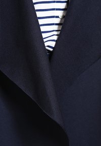 Blazer blu navy con un ampio rever, tessuto strutturato e una camicia a righe bianche e blu navy sotto.