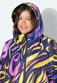 Femme portant une veste à capuche colorée avec des motifs abstraits jaunes, roses et violets, souriant légèrement, face à l'avant contre un fond uni.