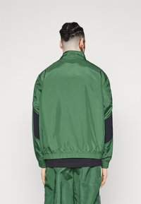 Chaqueta de satén verde con cuello alzado, puños y dobladillo elásticos. Presenta paneles laterales negros y una textura suave.