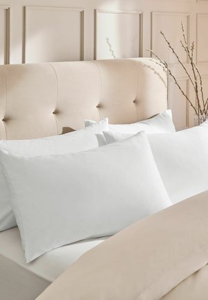 Beige gepolstertes Kopfteil mit vier weißen Kissen, die auf einem Bett mit beiger Bettwäsche arrangiert sind, und dekorative Zweige in einer Vase in der Nähe.