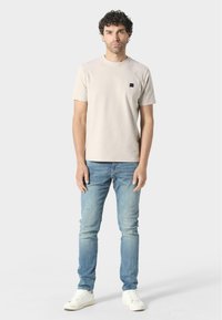 T-shirt in cotone beige chiaro con colletto girocollo e un piccolo logo nero sul lato sinistro del petto, abbinato a jeans blu chiaro e sneakers bianche.
