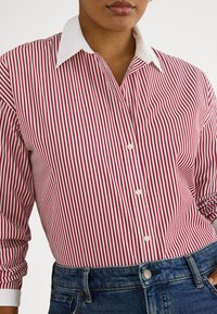 Άτομο που φοράει κόκκινο και άσπρο πουκάμισο με κάθετες ρίγες και λευκό γιακά, το πουκάμισο είναι μπα tucked μέσα σε μπλε τζιν τζιν με μπροστινό κουμπί.