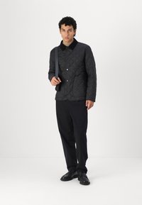 Casaco preto acolchoado com colarinho, botões de pressão e dois bolsos frontais. O modelo usa calças escuras e sapatos pretos, segurando uma alça.